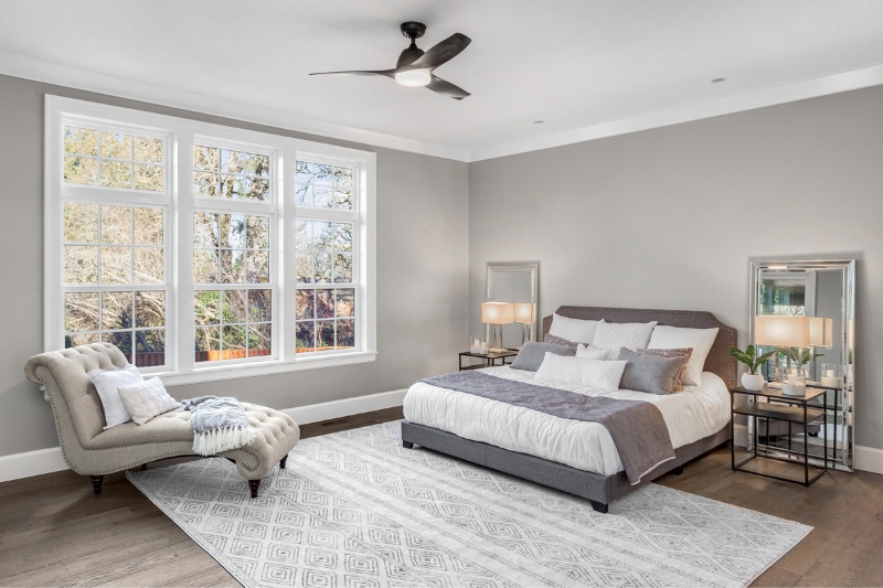 A modern bedroom with large windows, a gray upholstered bed with white and gray bedding, matching nightstands with lamps, a tufted chaise lounge, a patterned rug, and light gray walls with wood flooring.