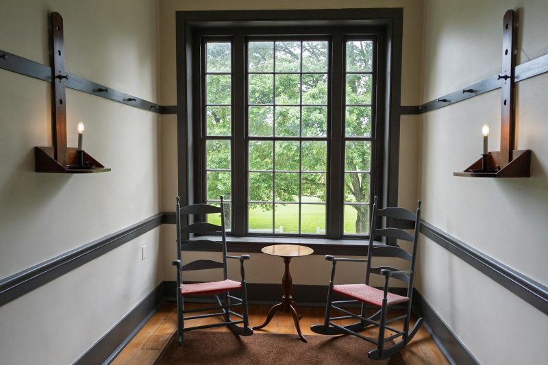 Cozy Shaker room with rocking chairs and candle sconces near a window, ideal for peaceful moments.