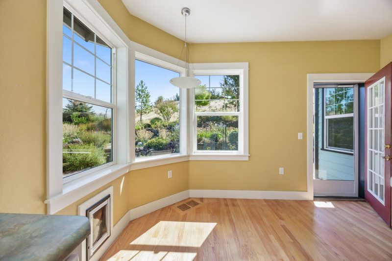 Spacious sunlit room with wooden floors, large windows, and views of a lush garden.