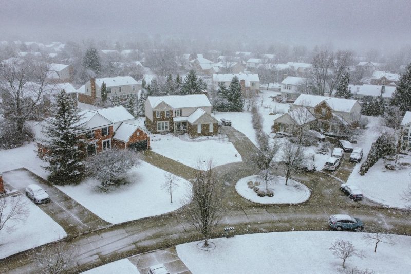 Aerial view of a snow-covered suburban neighborhood, featuring houses, trees, and quiet streets on a winter day.